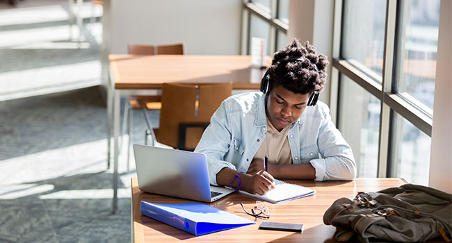 young male student with headphones taking notes in college library