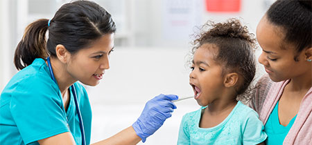 A nurse checks a little girl's mouth as she sits on her mother's lap