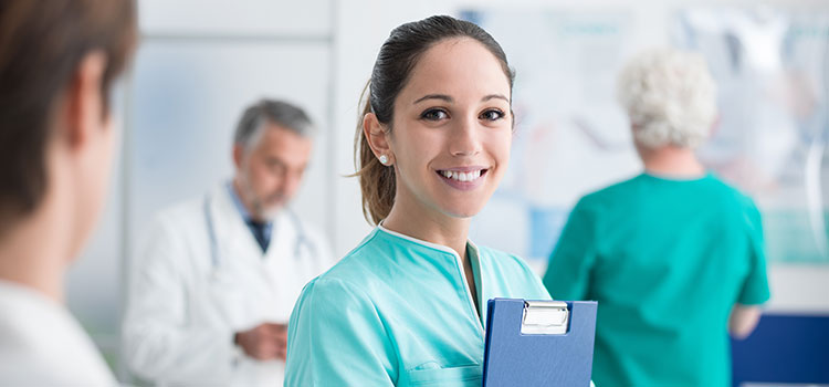 female nurse in teal scrubs smiles at camera while holding clipboard