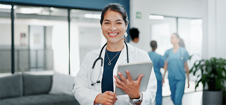 nurse with tablet and stethoscope around neck stands in busy hospital hallway
